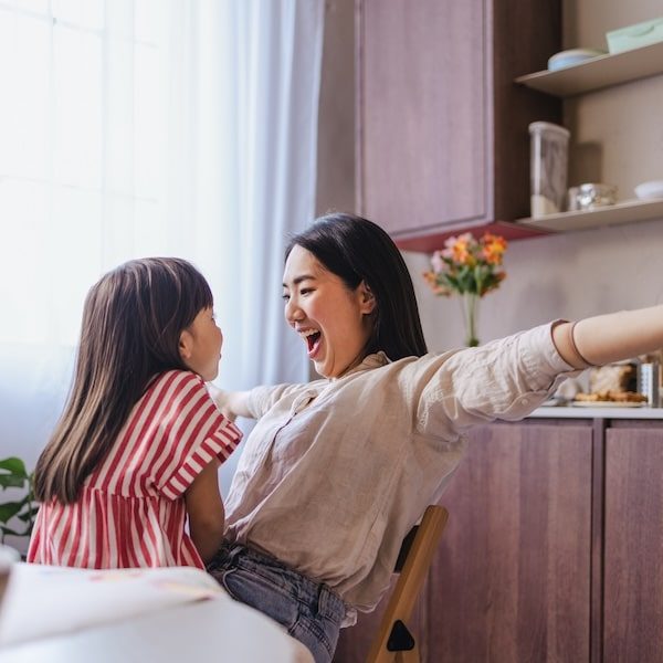 A wide angle view of an adorable playful Japanese child and her mother having fun while sitting in the kitchen.